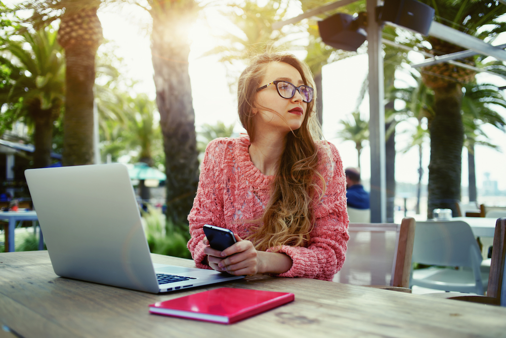 Young creative woman work on laptop while having breakfast on terrace, flare sun